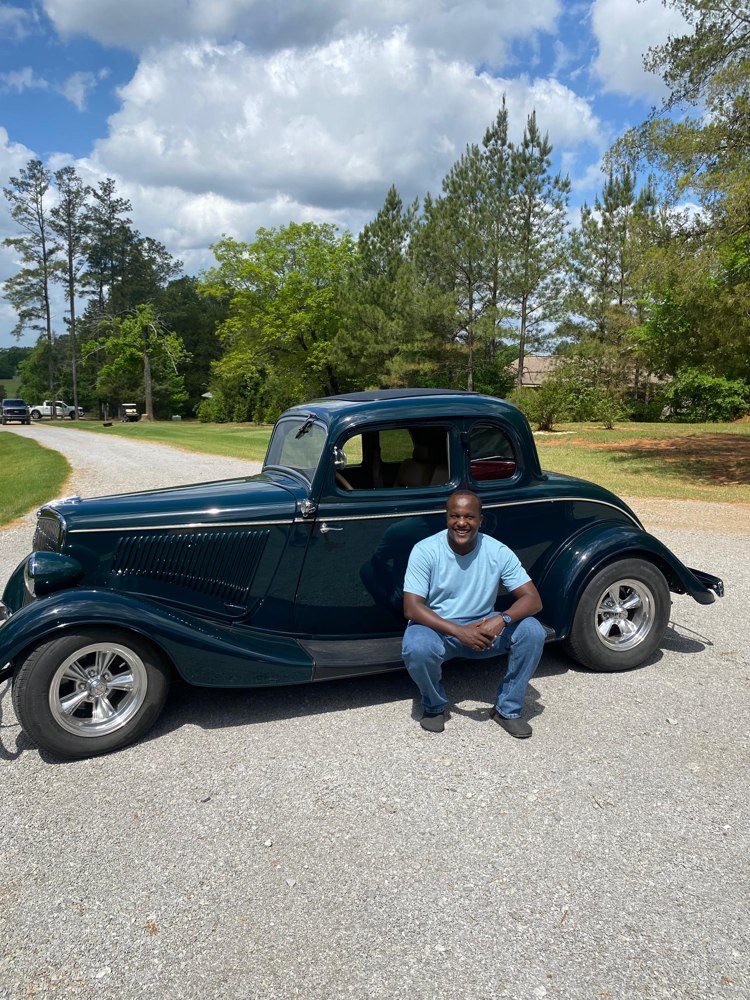 Enock Nkulanga crouching beside a vintage car in Mississippi